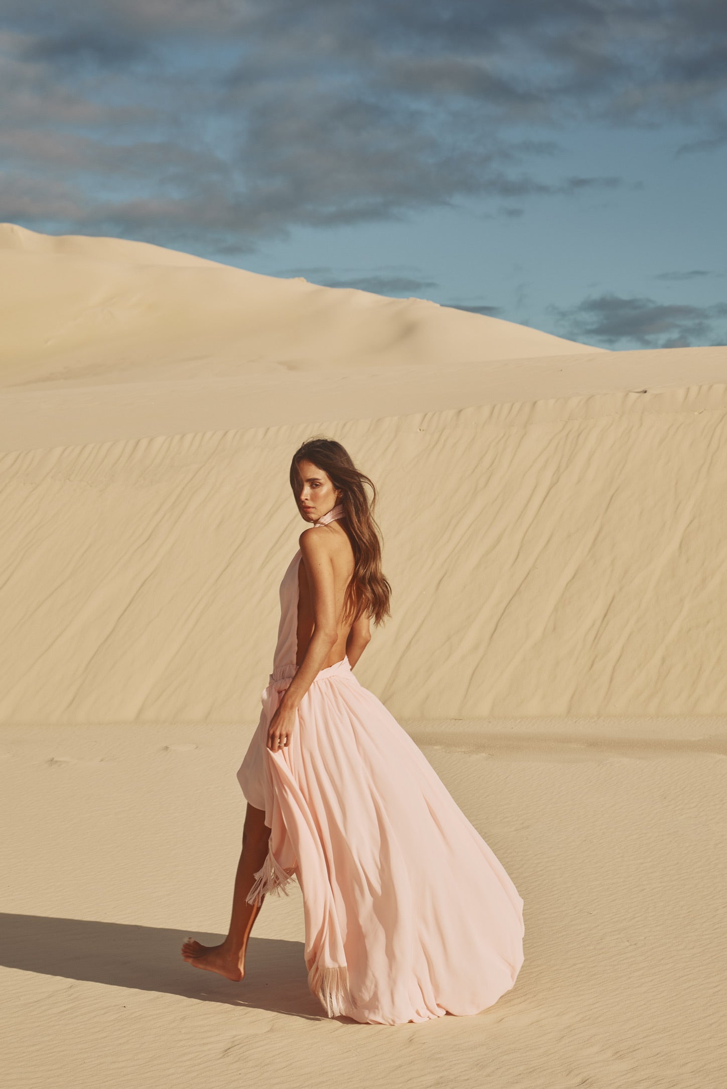 A woman in a flowing pale pink sleeveless dress walks barefoot across large sand dunes under a blue sky with scattered clouds.
